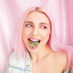 A woman with pink hair poses playfully with colorful candies in her mouth against a pink backdrop.