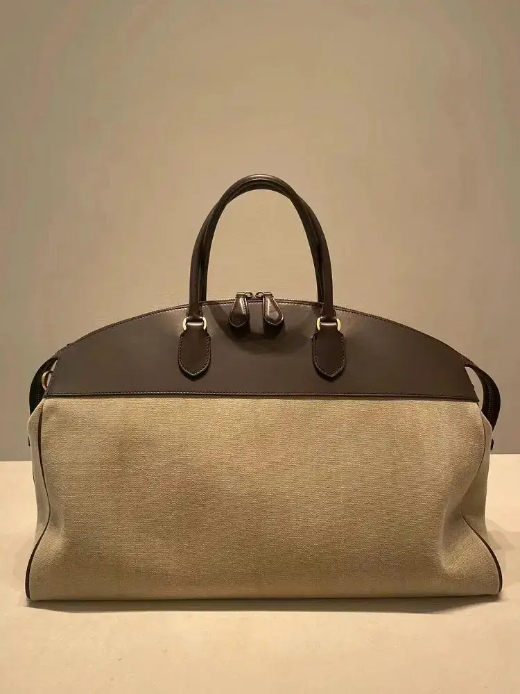 A beige canvas handbag with brown leather details. Beige canvas handbag with brown leather accents and handles displayed against a neutral background.
