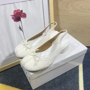 White slingback heels with a bow detail, placed on a white box next to a floral artwork.