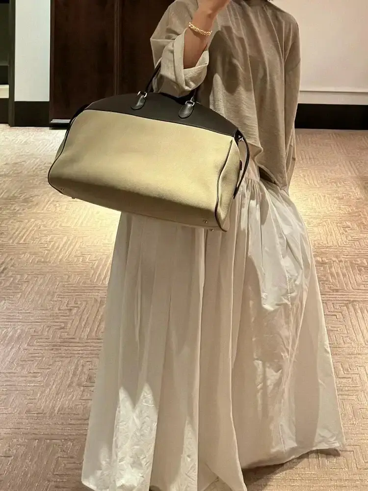 A handbag paired with a pleated skirt. A beige and black handbag held by a person wearing a long pleated white skirt and a gray top.