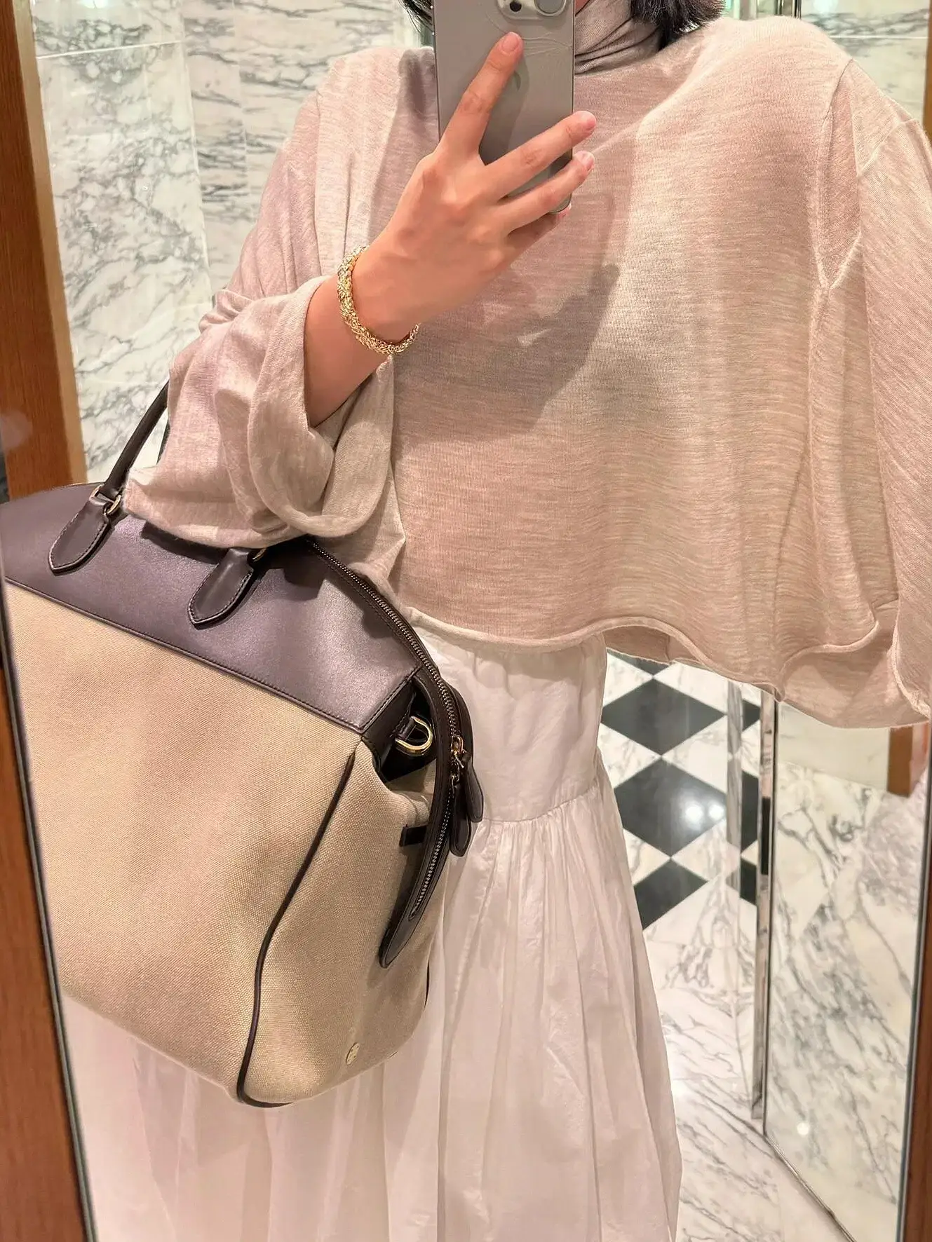 A woman taking a selfie while holding a handbag. A woman in a beige top holding a handbag while taking a selfie in a mirror.