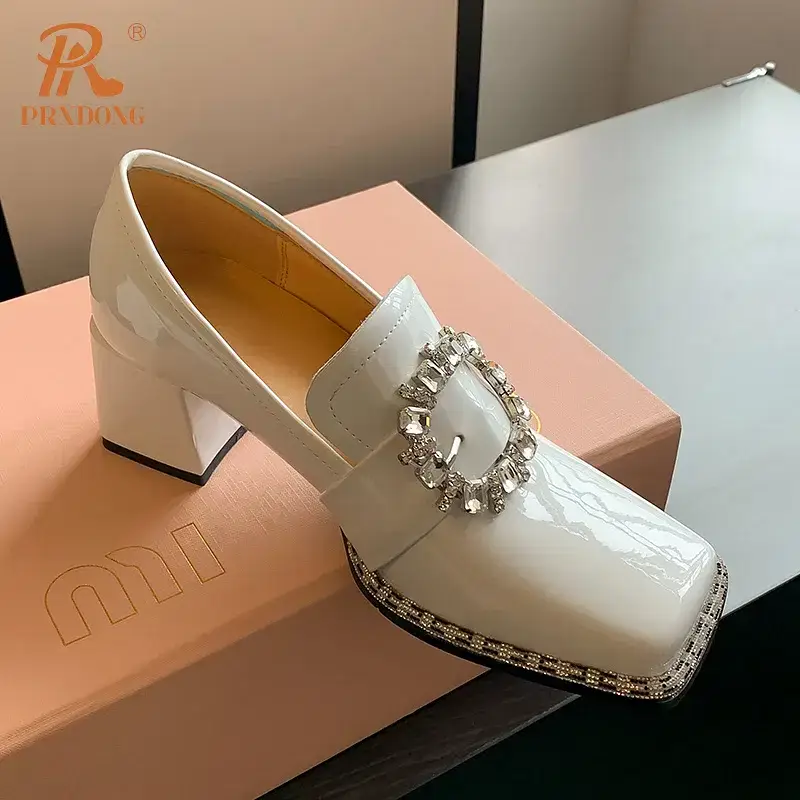 White patent leather shoes with a decorative buckle on the front. White patent leather heeled shoes featuring a decorative buckle and square toe design.