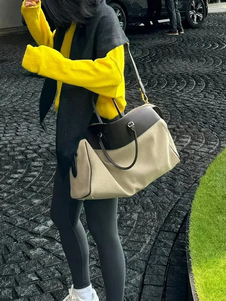 A woman dressed in a yellow sweater and gray scarf carrying a large handbag. Woman wearing a yellow sweater and gray scarf, holding a large beige and black handbag.
