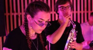 Two musicians performing together, one playing a saxophone and the other on a keyboard with pink lighting.