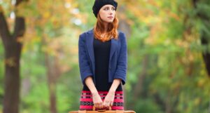 Woman with red hair wearing a black beanie and a blazer, holding a suitcase in an outdoor setting.
