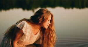 A woman with long hair in a floral dress leaning over the surface of a calm body of water.